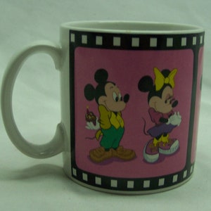 May include: White ceramic mug with a pink background and a black film strip border. The image features Mickey Mouse and Minnie Mouse in a cartoon style. Mickey is holding a small cake and Minnie is holding a present.