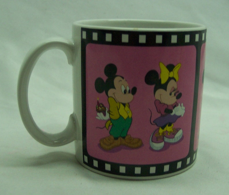 May include: White ceramic mug with a pink background and a black film strip border. The image features Mickey Mouse and Minnie Mouse in a cartoon style. Mickey is holding a small cake and Minnie is holding a present.