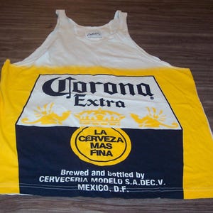 May include: A white tank top with a yellow and blue Corona Extra beer logo. The logo includes the text "Corona Extra", "La Cerveza Mas Fina", "Brewed and bottled by Cerveceria Modelo S.A.DEC.V. Mexico, D.F."