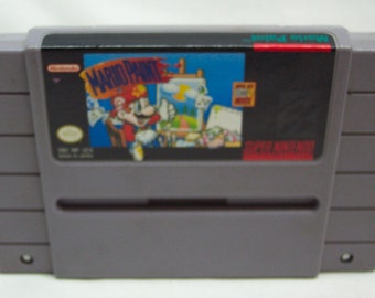 May include: A gray Super Nintendo Entertainment System (SNES) game cartridge with a black label. The label features a colorful image of Mario painting with a brush. The game title "Mario Paint" is printed in white letters on the label.