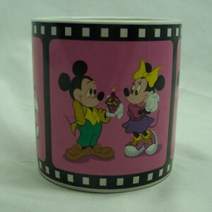 May include: A pink ceramic mug with a film strip border. Mickey Mouse and Minnie Mouse are depicted on the mug. Mickey is holding a cupcake with a single candle and Minnie is holding a cupcake.
