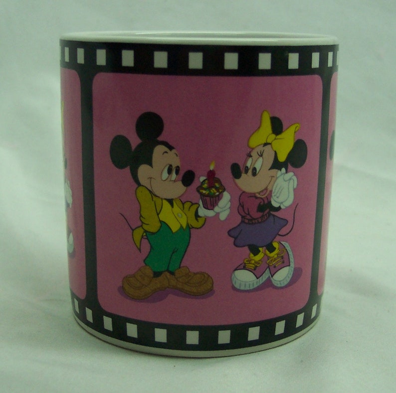 May include: A pink ceramic mug with a film strip border. Mickey Mouse and Minnie Mouse are depicted on the mug. Mickey is holding a cupcake with a single candle and Minnie is holding a cupcake.