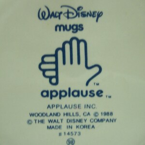 May include: A blue and white ceramic mug with the text "WALT Disney mugs" and "applause" with a trademark symbol. The mug also has the text "APPLAUSE INC. WOODLAND HILLS, CA &copy; 1988 THE WALT DISNEY COMPANY MADE IN KOREA #14573 (98)"