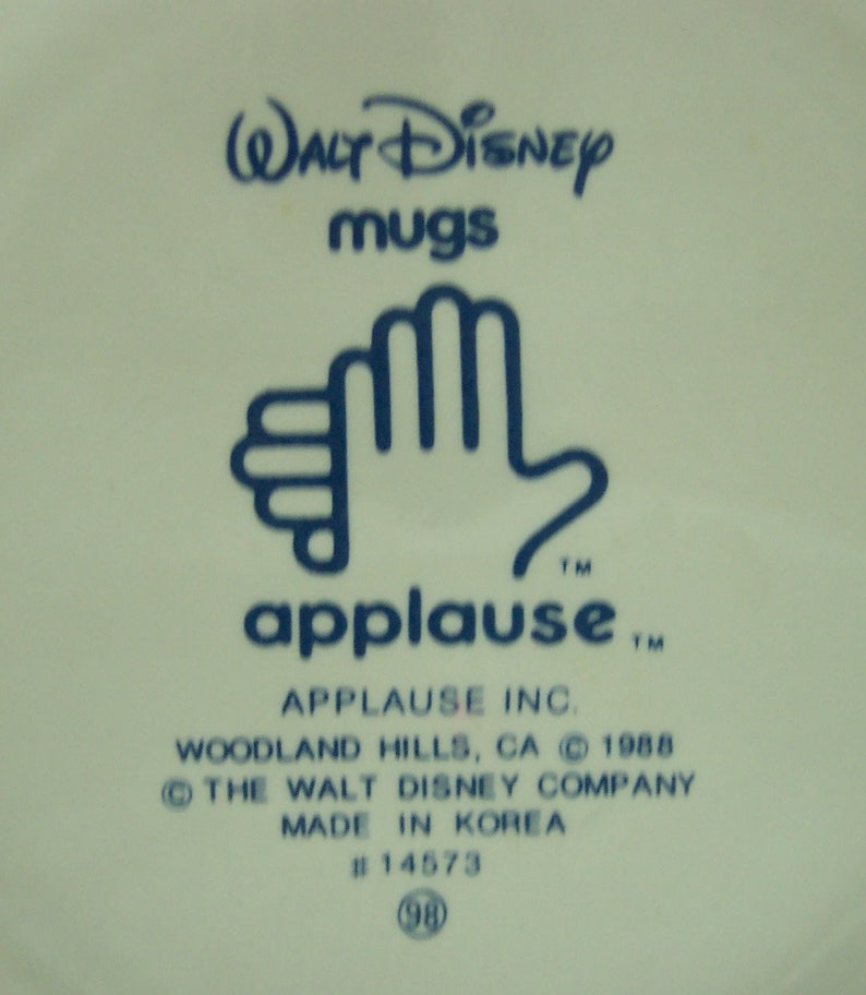 May include: A blue and white ceramic mug with the text "WALT Disney mugs" and "applause" with a trademark symbol. The mug also has the text "APPLAUSE INC. WOODLAND HILLS, CA &copy; 1988 THE WALT DISNEY COMPANY MADE IN KOREA #14573 (98)"