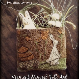 Może przedstawiać: Brązowa torba filcowa z haftowanym białym królikiem i koszykiem kwiatów. Torba jest wypełniona słomą i białymi kwiatami. Tekst "Spring Arrives On Widsbee Farm PN Pattern 001 2015" jest napisany na białej chmurze nad torbą. Tekst "Vermont Harvest Folk Art by Doreen Frost" jest napisany pod torbą.