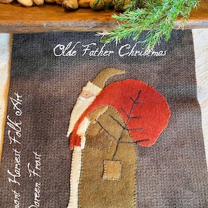 Può includere: Un arazzo da parete in feltro marrone con un'applique rossa, bianca e verde di una figura di Babbo Natale in piedi davanti a un albero. Il testo "Olde Father Christmas" è scritto in bianco nella parte superiore dell'arazzo. Il testo "Vermont Harvest Folk Art by Doreen Frost" è scritto in bianco sul lato sinistro dell'arazzo.