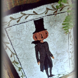 May include: A white needlepoint rug with a green border and a black and orange pumpkin figure wearing a top hat. The rug is titled "Vermont Harvest Folk Art Original Needlework Designs by Doreen Frost".