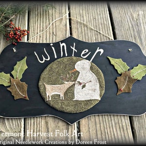 May include: A black wooden sign with the word "Winter" written in white paint. The sign features a felt appliqué of a snowman and a deer in a green circle, surrounded by felt holly leaves.
