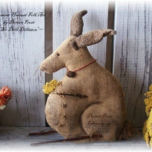 May include: A brown burlap bunny rabbit with a red ribbon around its neck. The bunny is holding a small bouquet of yellow flowers. The bunny is sitting on a wooden surface. The text "Permet Harvest Folk Art by Doreen Frost ~Sir Dill Dilliston~" is written on the surface behind the bunny. The text "Dereen Frost February" is written on the bunny's side.