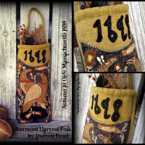May include: A brown felt wall hanging with a stitched design of two birds and the year 1698. The hanging has a loop at the top and a pocket on the front. The text "Autumn in Olde Massachusetts 1698" is written on the right side of the hanging. The text "Vermont Harvest Folk by Doreen Frost" is written at the bottom of the hanging.