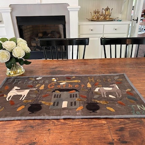 May include: A gray and brown fabric runner with the text "Friends Gather Here" and a design of a house, trees, and animals. The runner is on a wooden table with a vase of white flowers.