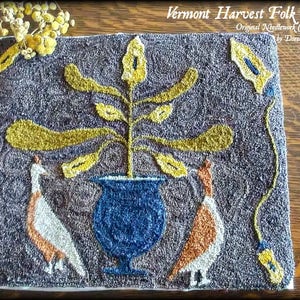May include: A needlepoint artwork featuring two birds positioned either side of a stylised plant in a blue vase. The background is a dark grey colour and the plant has yellow leaves. The artwork is titled "Vermont Harvest Folk Art" and is signed by Doreen Pratt.