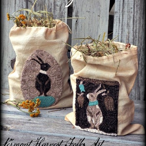 May include: Two cream-coloured canvas bags with embroidered bunny designs. The bags are filled with dried hay and have the text "Bunny Socks" written above them. The text "Vermont Harvest Folk Art by Doreen Frost" is written below the bags.