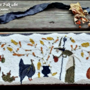 May include: A white needlepoint wall hanging with a folk art design featuring a witch stirring a cauldron, a squirrel, two turkeys, and a pumpkin. The design is surrounded by a brown and orange border. The text "Vermont Harvest Folk Art" is at the top of the piece.