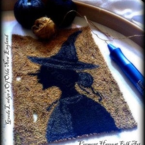 May include: A black silhouette of a witch with a pointed hat and long hair, stitched on a brown fabric background. The fabric is in a hoop and there are balls of yarn and a needle nearby. The text "Vermont Harvest Folk Art by Doreen Frost" is visible below the image.
