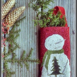 May include: A red felt stocking with a white snowman holding a green Christmas tree. The stocking has a green felt trim and the text "When Snowflakes Fall" is printed at the bottom.
