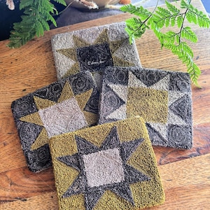 May include: Four square, textured coasters with a starburst design. The coasters feature a central square surrounded by star points in shades of gold, gray, and black. They are arranged on a wooden surface, with green foliage in the background.