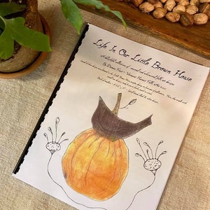 May include: A book titled "Life In Our Little Brown House" with a hand-drawn illustration of a pumpkin with a bird on top and two small figures with outstretched arms on either side.
