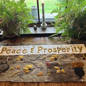 May include: A handcrafted textile table runner with the words "Peace & Prosperity" embroidered in gold. The runner features a turkey, flowers, and a vase design in shades of brown, yellow, and green. Two potted ferns frame the scene.