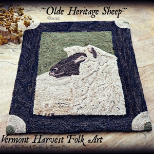Op de afbeelding: Een handgemaakt tapijt met een schapenmotief. Het tapijt heeft een donkere rand met lichtgekleurde hoekaccenten. Het schaap heeft een zwart gezicht en beige wol. De tekst "Olde Heritage Sheep" en "Vermont Harvest Folk Art" zijn zichtbaar.