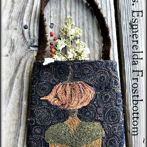 May include: A small, black and brown fabric bag with a pumpkin-shaped design and a brown fabric handle. The bag is decorated with a green, yellow, and orange pumpkin with a brown stem. The bag is hanging on a wooden surface. "Vermont Harvest Folk Art Original Needlework Creations by Doreen Frost" is written below the bag.