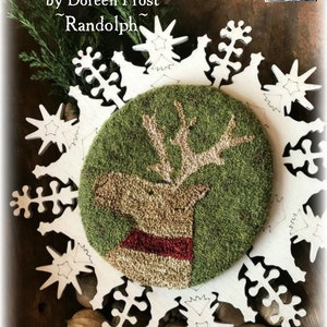 May include: A white wooden snowflake frame with a green and brown needlepoint design of a reindeer inside. The needlepoint design is surrounded by a green background. The text "Vermont Harvest Folk Art" is at the top of the image. The text "Original Needlework Creations" is below the first line of text. The text "by Doreen Frost" is below the second line of text. The text "~Randolph~" is below the third line of text.