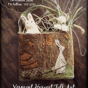 May include: A brown felt bag with a white bunny rabbit and a basket of flowers embroidered on it. The bag is filled with dried grass and white flowers. The text "Spring Arrives On Widsbee Farm PN Pattern 001 2015" is written in a white cloud shape above the bag. The text "Vermont Harvest Folk Art by Doreen Frost" is written below the bag.