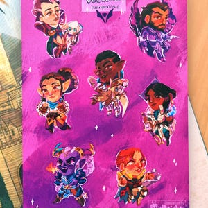 May include: A set of eight colourful stickers featuring characters from the VeilGuard campaign. The stickers are on a pink background with glitter accents. The text "VeilGuard Campaign" is at the top of the sheet.