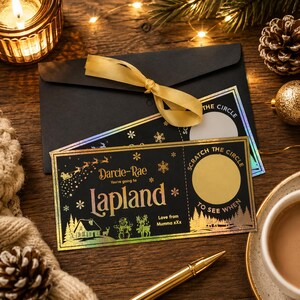 May include: A black gift card with gold foil detailing, featuring the text "Lapland" and "Darcie-Rae". The card has a scratch-off area and is tied with a gold ribbon. A black envelope and a lit candle are in the background.