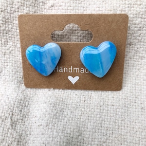 Op de afbeelding: Een paar blauwe en witte hartvormige oorbellen op een bruine kaart met het woord "Handmade" erop.