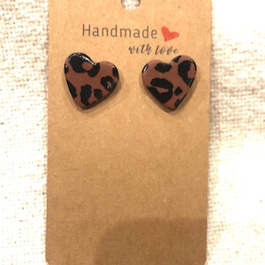 Peut inclure: Une paire de boucles d'oreilles en forme de cœur brun avec un motif léopard noir. Les boucles d'oreilles sont sur une carte brune avec le texte "Handmade with love" en rouge et blanc.