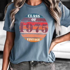May include: A blue short-sleeved t-shirt with a vintage-style graphic of a red, orange, and yellow sunburst with the text "Class of 1975 Vintage" printed on it.
