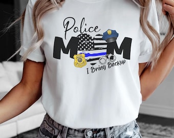 May include: White t-shirt with a graphic design featuring the words "Police Mom" above an American flag heart, police badge, handcuffs, and a police hat. The text "I Bring Backup" is below the design.