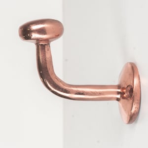 May include: A copper-colored, curved handrail bracket. The bracket has a rounded top, a curved arm, and a circular base with a single mounting hole. The metal has a polished finish, reflecting light. The bracket is designed for wall mounting.