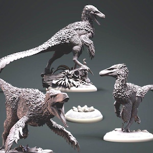 Op de afbeelding: Drie gedetailleerde, grijze, gevederde dinosaurusfiguren op aparte bases. De dinosaurussen staan in verschillende dynamische poses, een van hen houdt een nest met eieren vast.