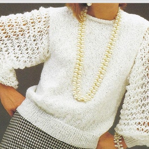 May include: White knitted sweater with a round neckline and long sleeves. The sleeves are made with a lacy stitch pattern. The sweater is worn over a black and white houndstooth skirt.
