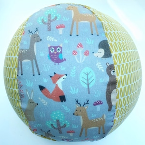 Puede incluir: Una pelota redonda de tela con estampado de animales del bosque. La pelota tiene un fondo azul claro con ilustraciones de ciervos, zorros, búhos y setas. Un lado tiene un patrón geométrico amarillo.