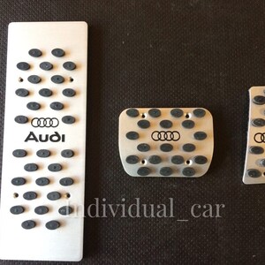 May include: Set of three silver-colored car pedal covers. Each pedal cover has a series of small, black, oval-shaped rubber grips. The Audi logo and brand name are printed in black on the largest pedal cover.