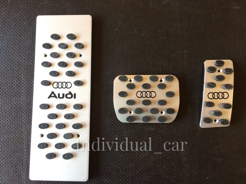 May include: Set of three silver-colored car pedal covers. Each pedal cover has a series of small, black, oval-shaped rubber grips. The Audi logo and brand name are printed in black on the largest pedal cover.