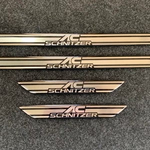 Könnte beinhalten: Vier silberne Metall-Einstiegsleisten für Autotüren mit dem Schriftzug "AC Schnitzer" in schwarzer Schrift.