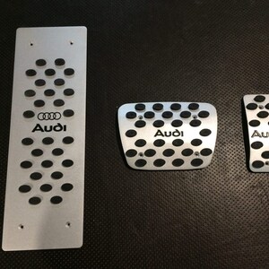 May include: Set of three silver-colored Audi car pedal covers. The covers have a perforated design with black rubber inserts and the Audi logo. The set includes a long footrest, a square pedal cover, and a smaller pedal cover.