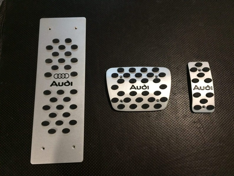 May include: Set of three silver-colored Audi car pedal covers. The covers have a perforated design with black rubber inserts and the Audi logo. The set includes a long footrest, a square pedal cover, and a smaller pedal cover.