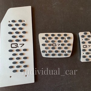 May include: Set of three silver-colored car pedal covers with black rubber grips. Each pedal cover has the text "Q7" printed in black. The covers are arranged on a dark surface.