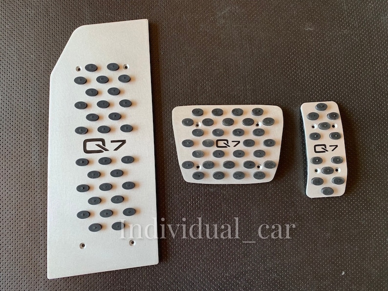 May include: Set of three silver-colored car pedal covers with black rubber grips. Each pedal cover has the text "Q7" printed in black. The covers are arranged on a dark surface.