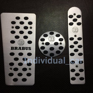 Puede incluir: Un juego de tres cubiertas de pedal de coche plateadas con inserciones de goma negra. Los pedales presentan el logotipo de BRABUS y una "B" negra en un círculo.