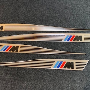 May include: Four silver metal car door trim pieces with a black and white striped pattern and a blue, red, and black BMW M logo.