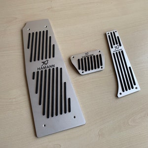 Puede incluir: Tres cubiertas de pedal de coche de metal plateado con inserciones de goma negra. Cada cubierta tiene la palabra "HAMANN" impresa en ella.
