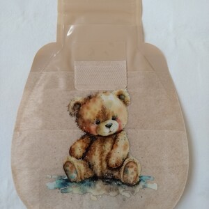 May include: A beige ostomy pouch with a brown teddy bear illustration. The pouch has a clear plastic flap and a beige adhesive strip.