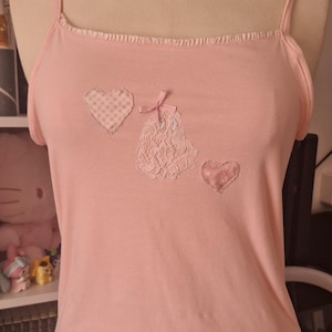 Peut inclure: Un caraco rose clair doté de fines bretelles et d'une encolure à volants. Le devant est orné de trois appliqués : un cœur à carreaux, un cœur en dentelle et un petit nœud rose, ainsi qu'un écusson en forme de cœur rose uni.