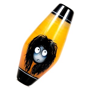 Puede incluir: Un tapón de botella decorativo con una ilustración de dibujos animados de un personaje de aspecto triste con cabello negro y ojos grandes. El tapón tiene una parte superior e inferior negras con un cuerpo amarillo y naranja.